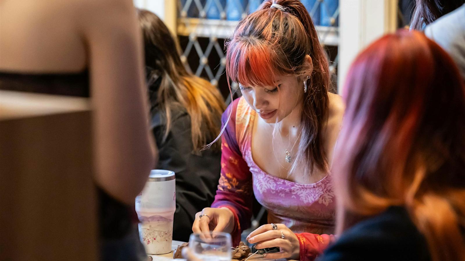 A person with long hair in a pony tail is sitting at a table and working with some clay.