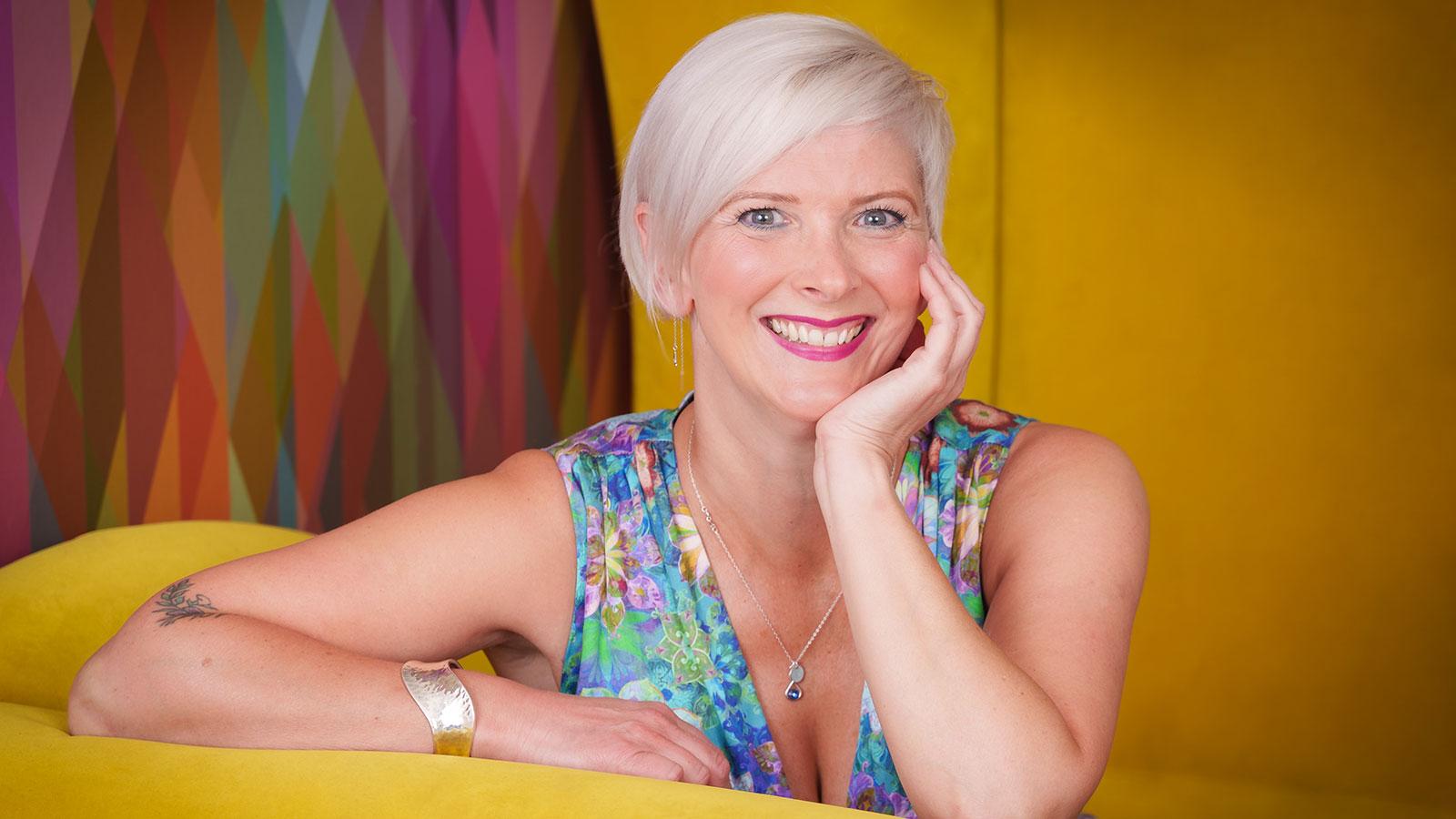 Opera singer Emma Morwood is sitting in a yellow room with a multicoloured piece of artwork behind her. She has short blond hair, wears a blue floral sleeveless dress and smiles to the camera.