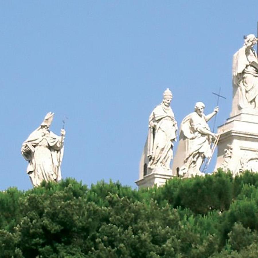 A series of white statues of people on a hill with some bushes and part of a building seen in the foreground