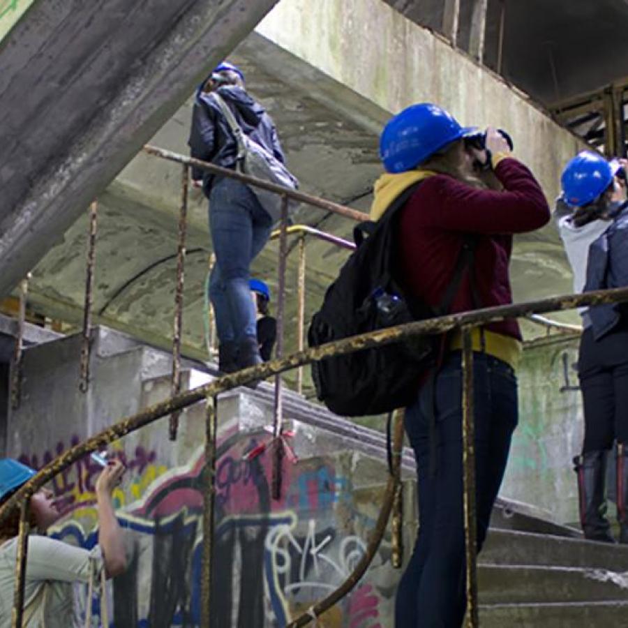 Three students on a spiral staircase are wearing blue hard-hats and taking photos of their surroundings, which is the internal of a building that looks to be in disrepair