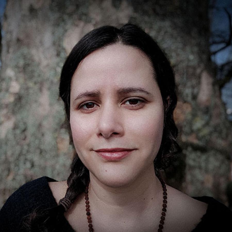 A person with dark hair, plaited, wearing a dark top and beaded necklace.