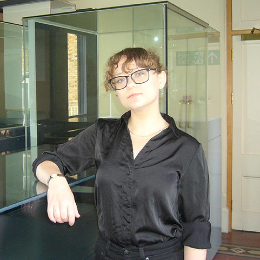 A person with light curly hair, wearing a black silk shirt and glasses, looks to the camera