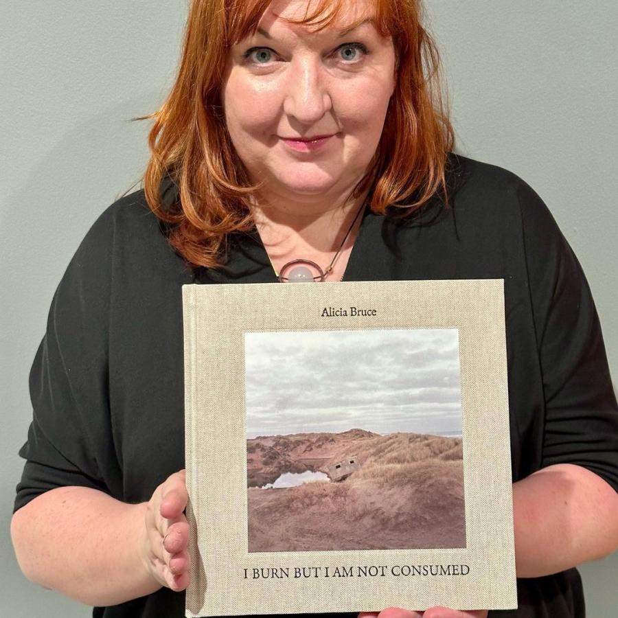 A person with short red hair looks at the camera while holding a book with a photo of sand dunes on it.