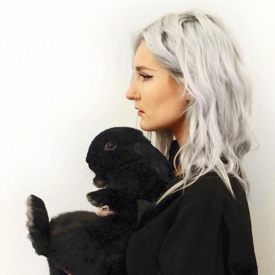 A person with long grey hair, standing to the side and holding a black rabbit in their arms.