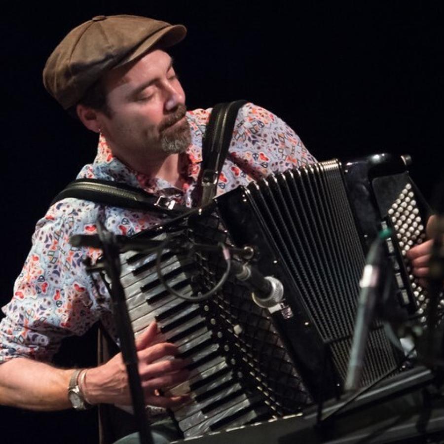 A person wearing a flat cap and patterned shirt, playing an accordion.