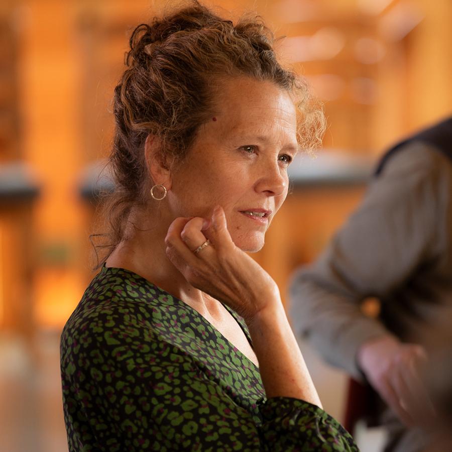 A photo of a person in a green patterned dress, wearing their hair up and bringing their hand to the side of their face.