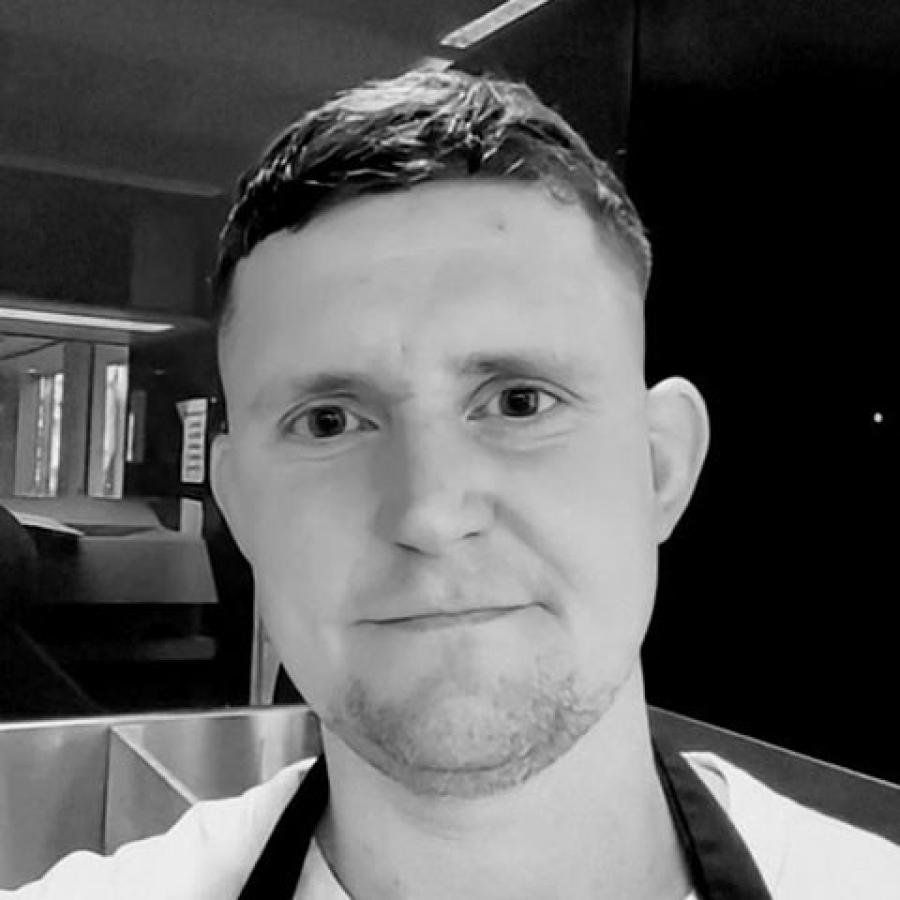 A black and white image of a person with short hair, wearing a white top and smiling at the camera.