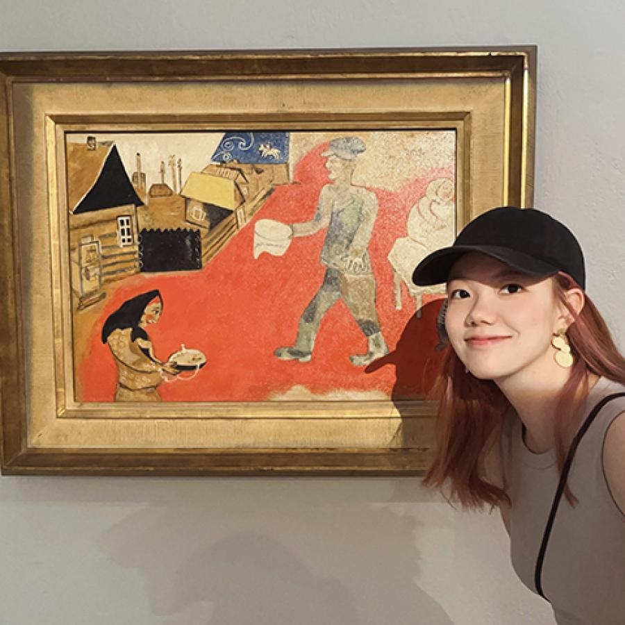 A person wearing a black cap and light top, standing next to a painting and smiling.