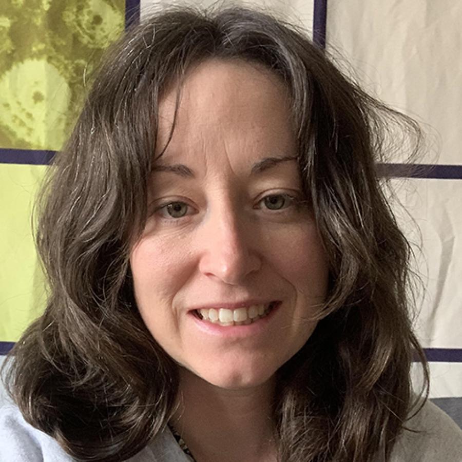 A person with long dark hair, wearing a white top and smiling to the camera.