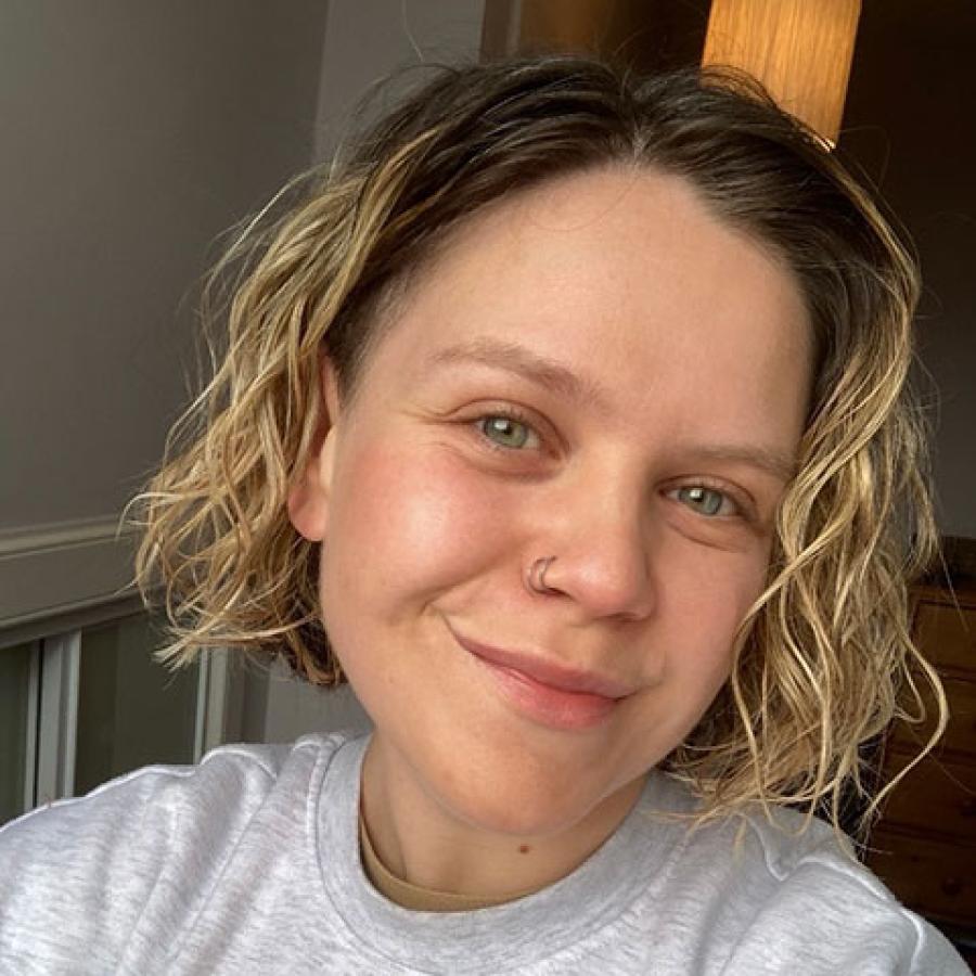 A person with short, blond curly hair, wearing a grey tshirt and smiling to camera.