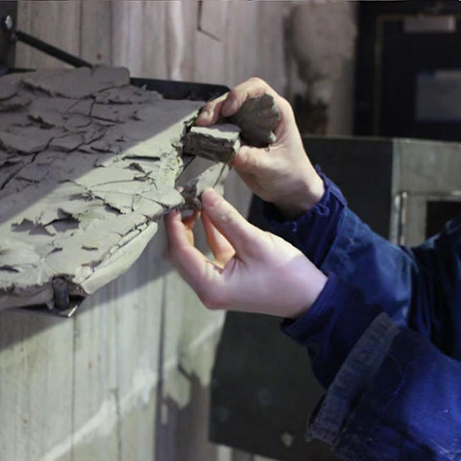 A pair of hands working with a sheet of broken clay that's perched on a metal shelf.
