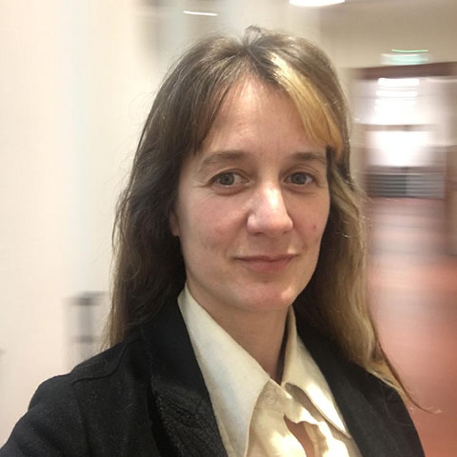 A person with long dark hair, with a chunk of blonde at the front of it, wearing a light shirt and black jacket. They are standing in one of ECA's hallways.