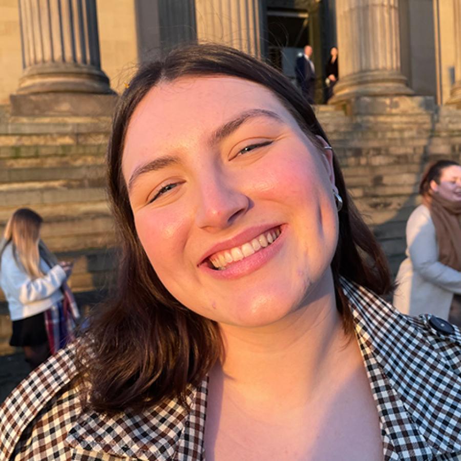 A person with short dark hair, smiles at the camera whilst standing in front of the steps and grand columns of a building.