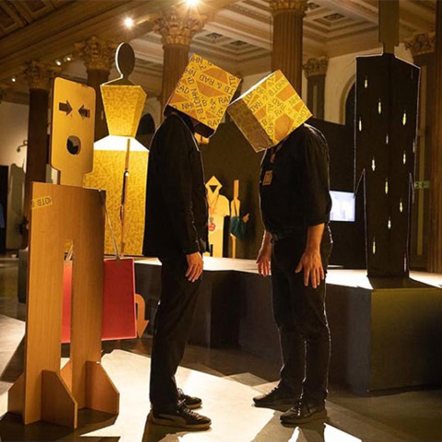 Two people standing in the Gallery of Modern Art, Glasgow, wearing boxes on their heads.