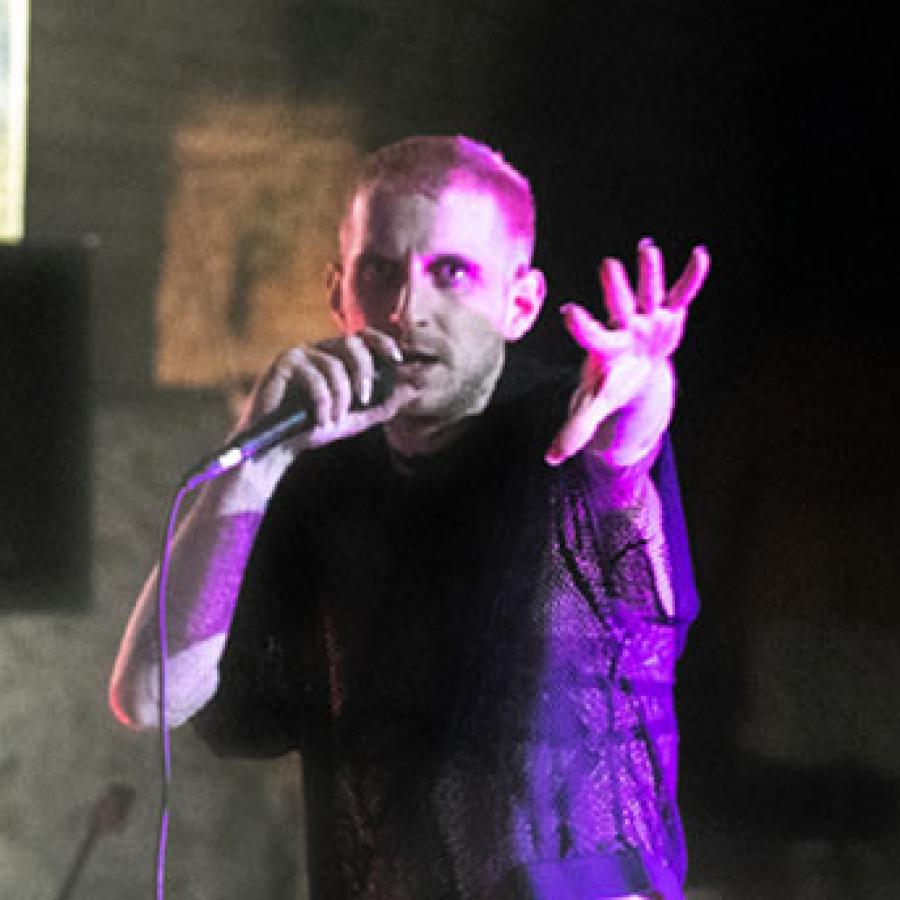 A person in a mesh top, holding a mic and reaching towards the camera.
