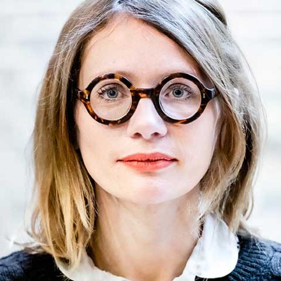 A person with shoulder length, lightbrown/blond hair, wearing round glasses and a white shirt under a blue cable knit jumper.