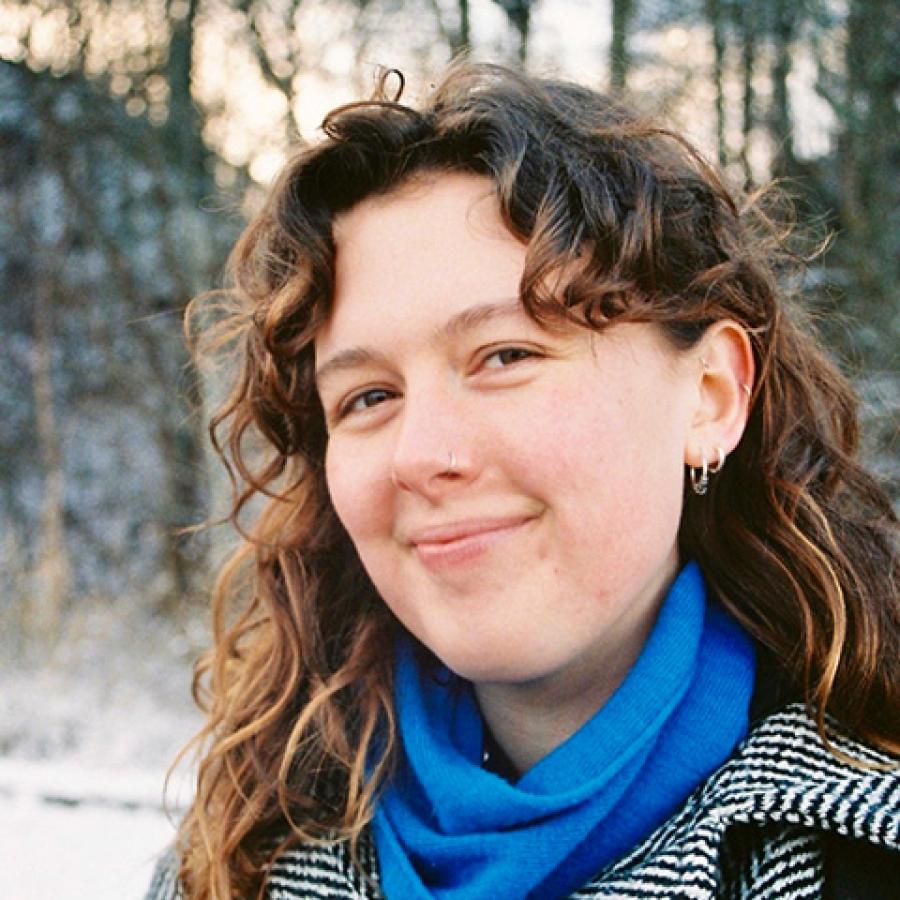 A person with long, light brown curly hair, wearing a blue scarf and grey herringbone coat. They are smiling at the camera.