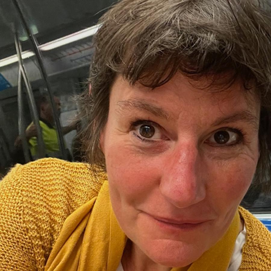 A person with short, dark brown hair, wearing a yellow knitted top, looks to the camera.