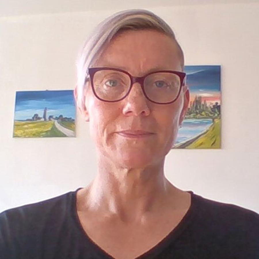 A person with short, silvery hair, wearing pink glasses and a dark top. They are looking directly to the camera, and have two paintings of landscapes hung up on the wall behind them.