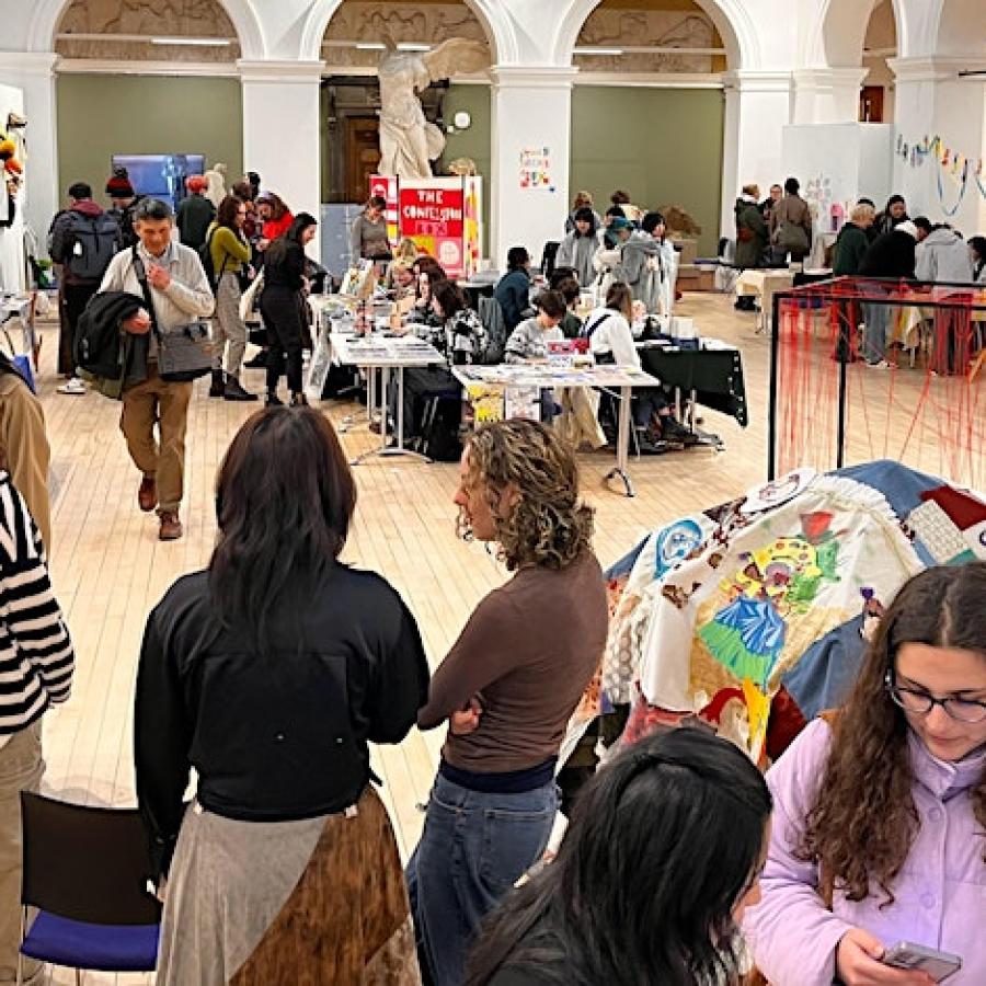 ECA's sculpture court set up with stalls for the annual winter fair. Students and members of the public mill about in the space looking at artwork, zines, prints and much more.