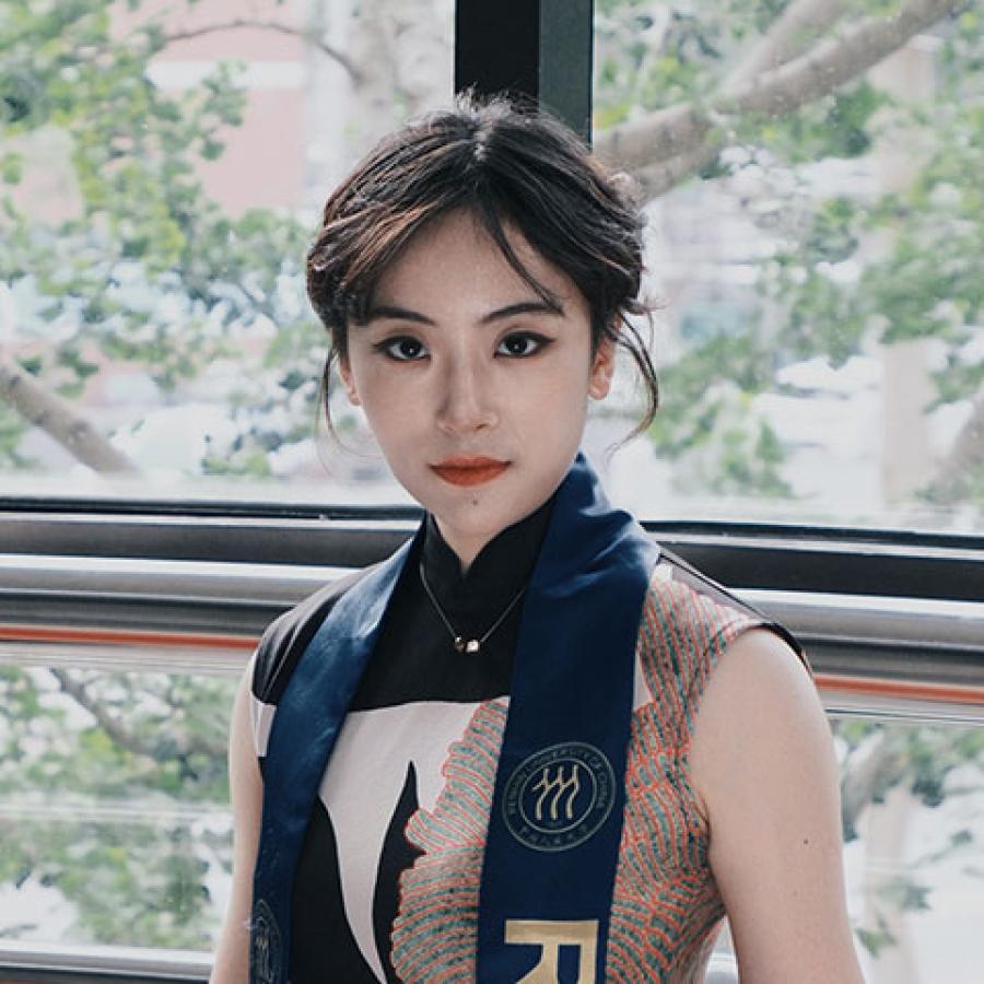 A person stands in front of a window looking out onto trees. They wear a dress and a blue sash draped around their neck. They're looking directly to camera.