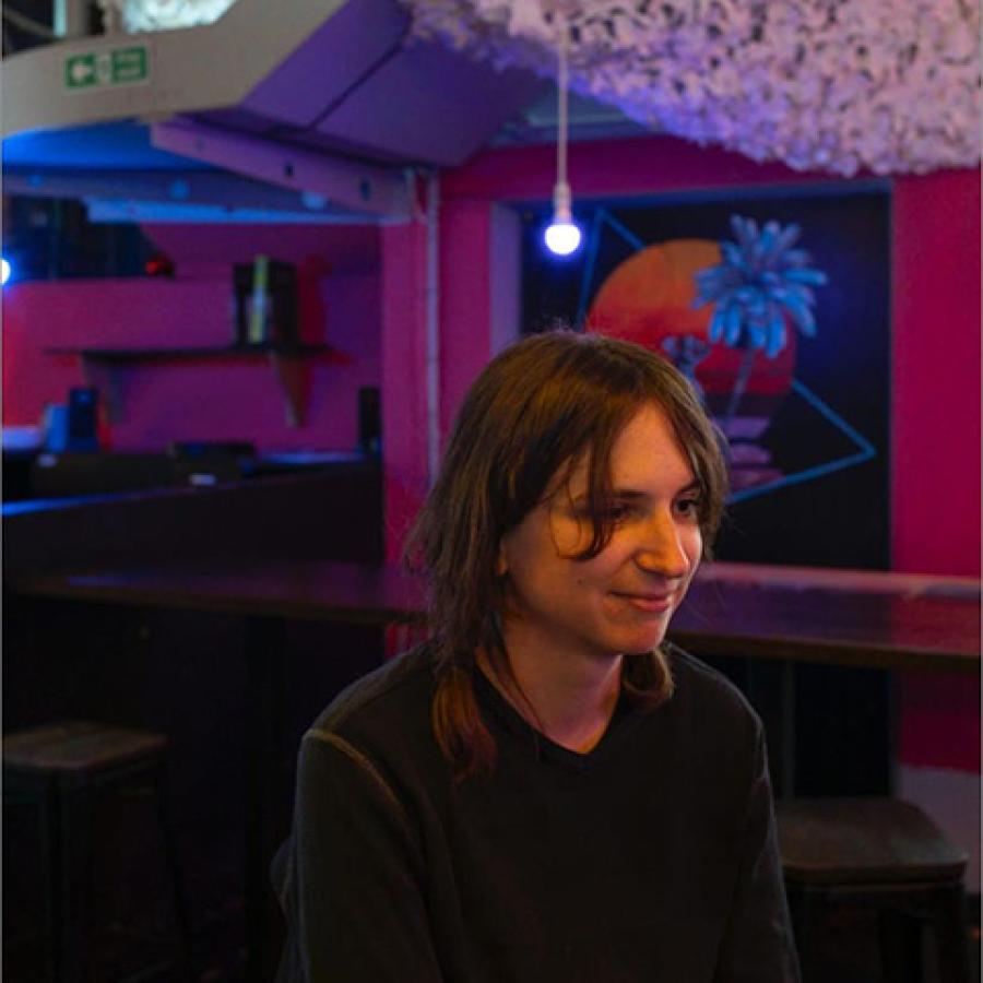 A person with dark hair, sits in a room with red walls. There is a bare lightbulb in the background and a white, decorative hanging on the ceiling.