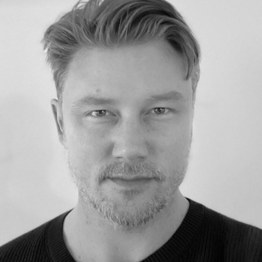 A black and white photo of a person with short light hair and they're wearing a black tshirt. They're looking directly into the camera.
