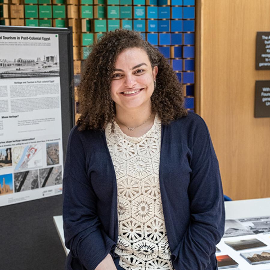 A person with dark brown, curly hair, is smiling to the camera. They wear a navy blue cardigan and cream lace top.