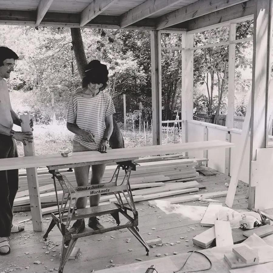A black and white photo of two people on a building site of a house. One is sawing wood, the other watches. 