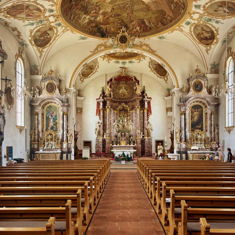 An image of the interior of the Heiligkreuzkirche in Kirchberg taken from the back of the church looking towards the altar. There are rows of wooden pews under the domed ceiling that's adorned with a fresco and gold, ornate cornicing and decoration.