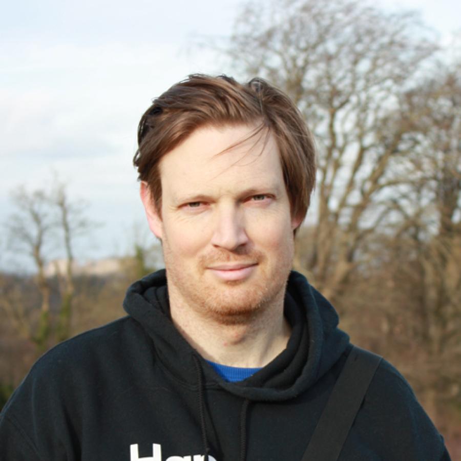 Dr Tom Mudd stands outside in a black hooded jumper. There are trees in the background.