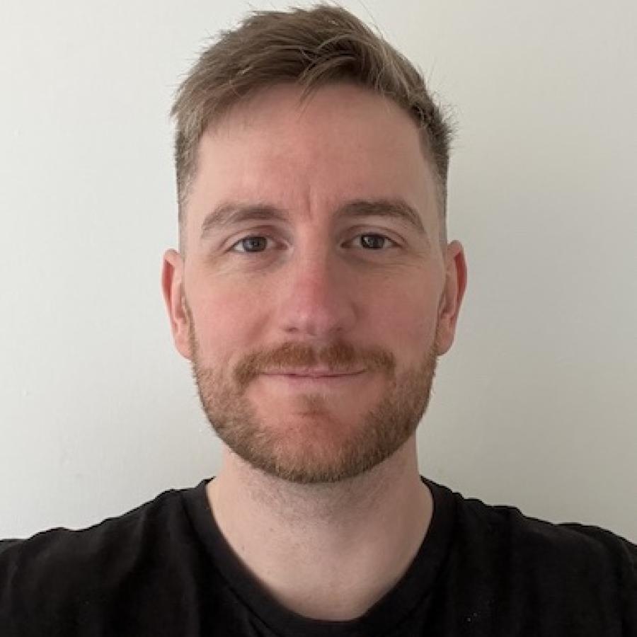 A person with sandy coloured hair, smiles to the camera. They're wearing a black tshirt and stand in front of a white wall.