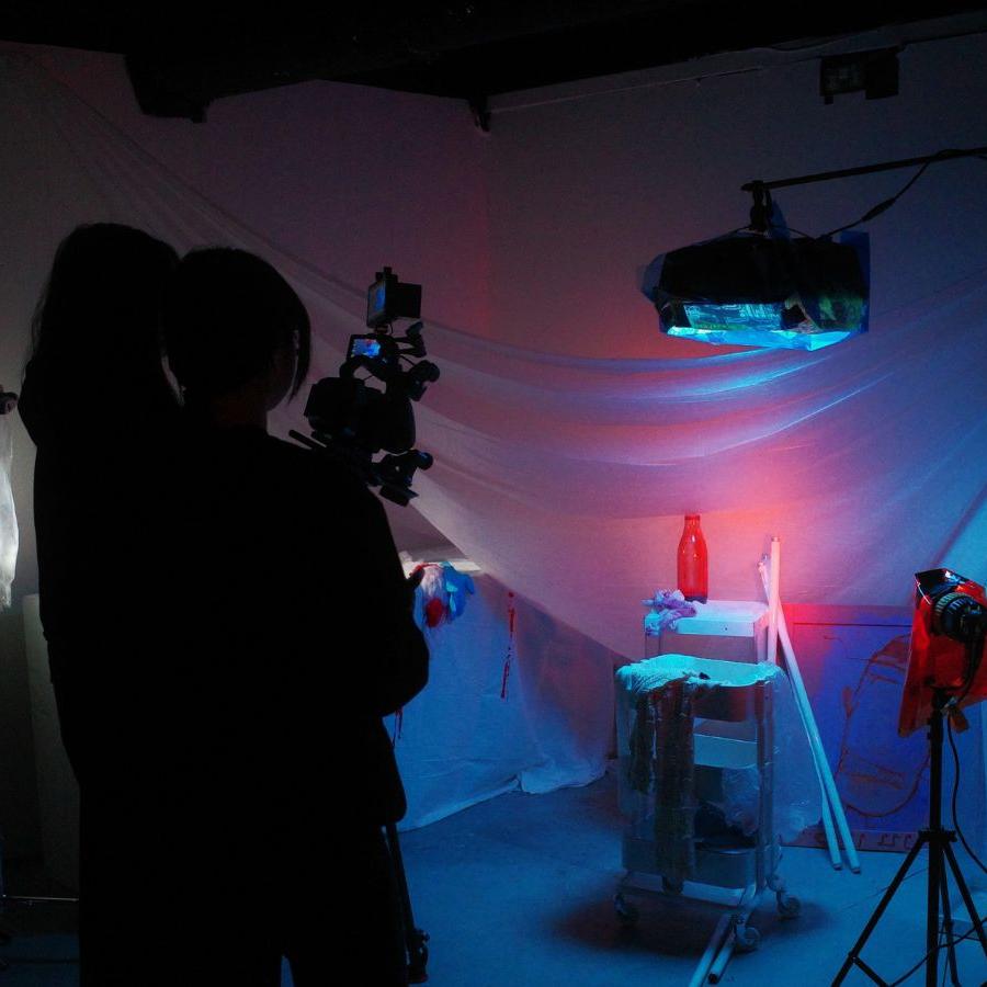 Three students are standing behind a camera, filing a low lit scene which includes a sheet hung up, a set of drawers on wheels and random objects like cloths, bottles, sticks etc littered about.