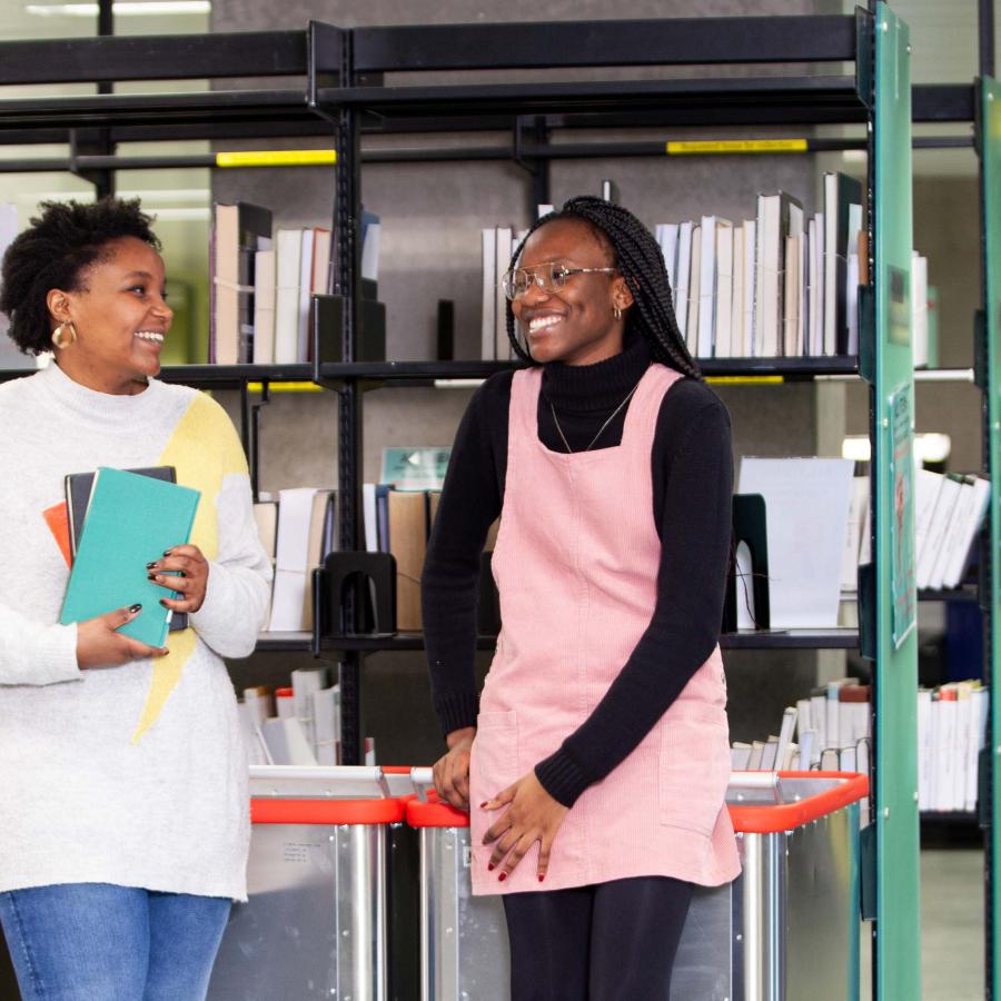 Three female students chatting and smiling in a library, with another female student walking in the background