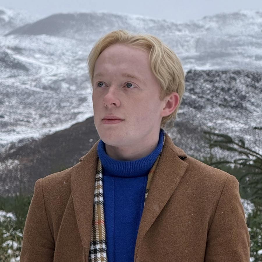 A person standing against a backdrop of snowy mountains. They wear a blue jumper and tan coat.