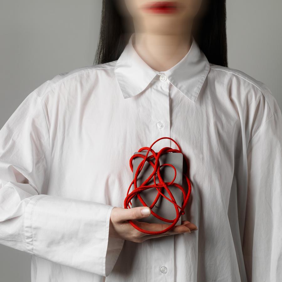 Person wearing a white shirt holds object to chest. The object is a grey box wrapped in a tangle of red cord. 