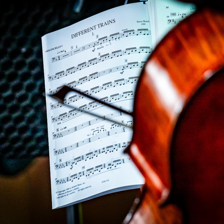 A cello with a bow held in front of a sheet of music called Different Trains.