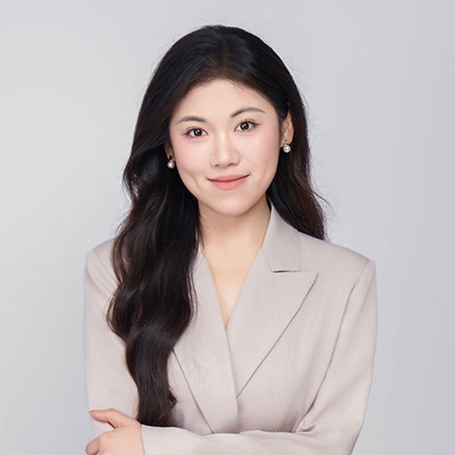 A person with long dark hair, wearing a beige suit jacket, smiles to the camera.