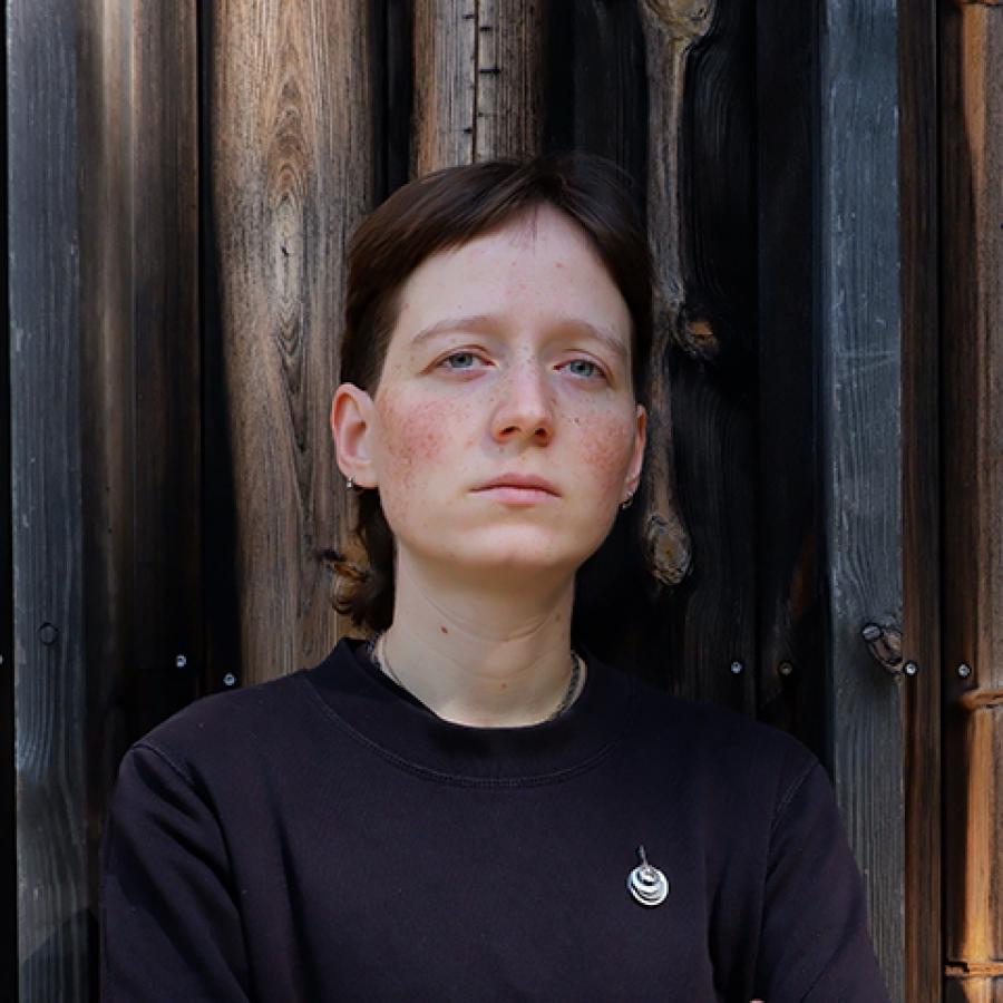 A person with short hair, wearing a navy blue jumper, stands in front of a what looks like a wooden cabin wall.