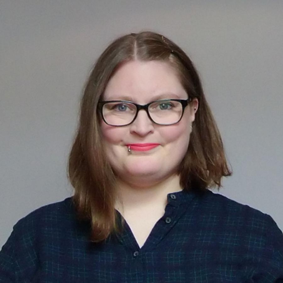 Suzanne black looks straight to camera. They have short, mid brown hair and wear black glasses and a black shirt.