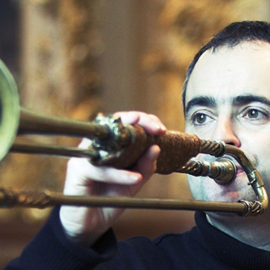 A person with short hair and wearing a dark shirt, blowing into a trombone.