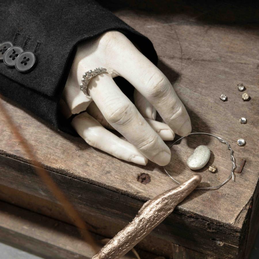 A sculpture of a hand with the third finger broken off. The hand is resting on a piece of wood, with small pieces of metal around it. 