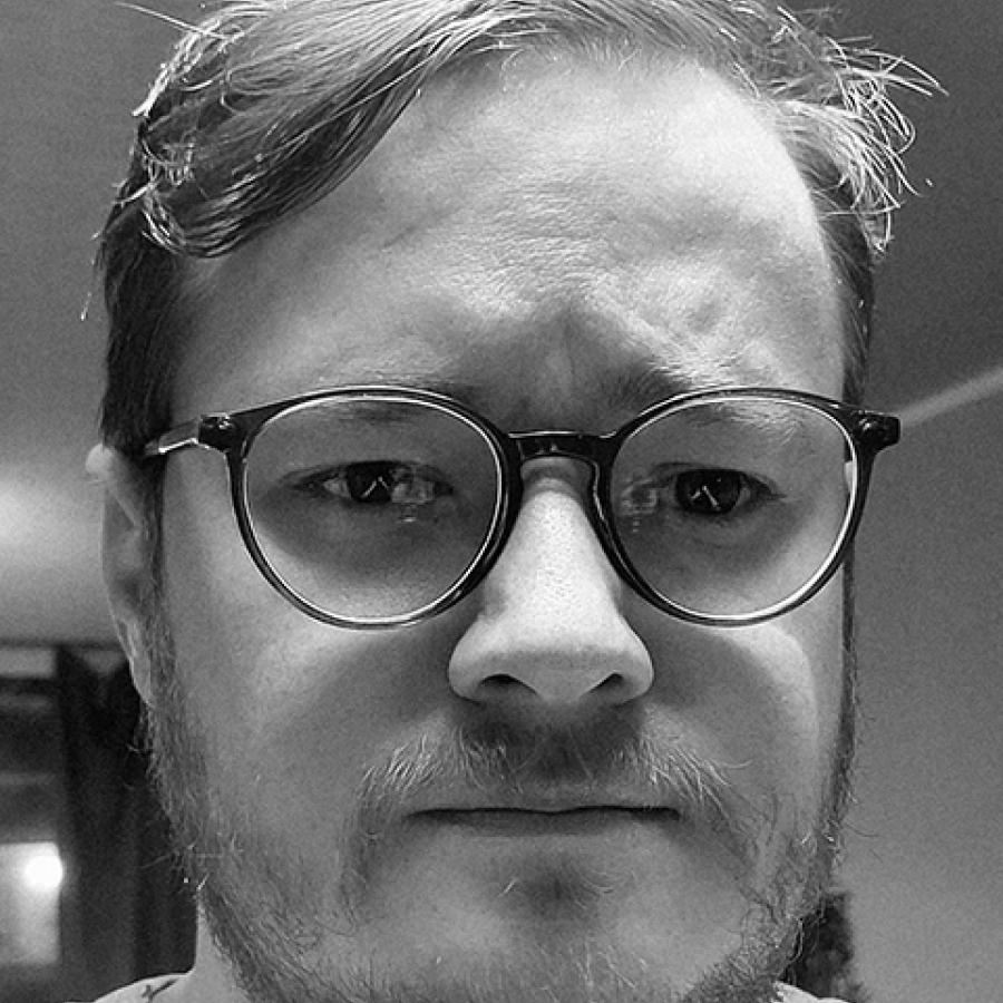 A black and white image of Dean Chalmers, looking to the camera and wearing glasses.