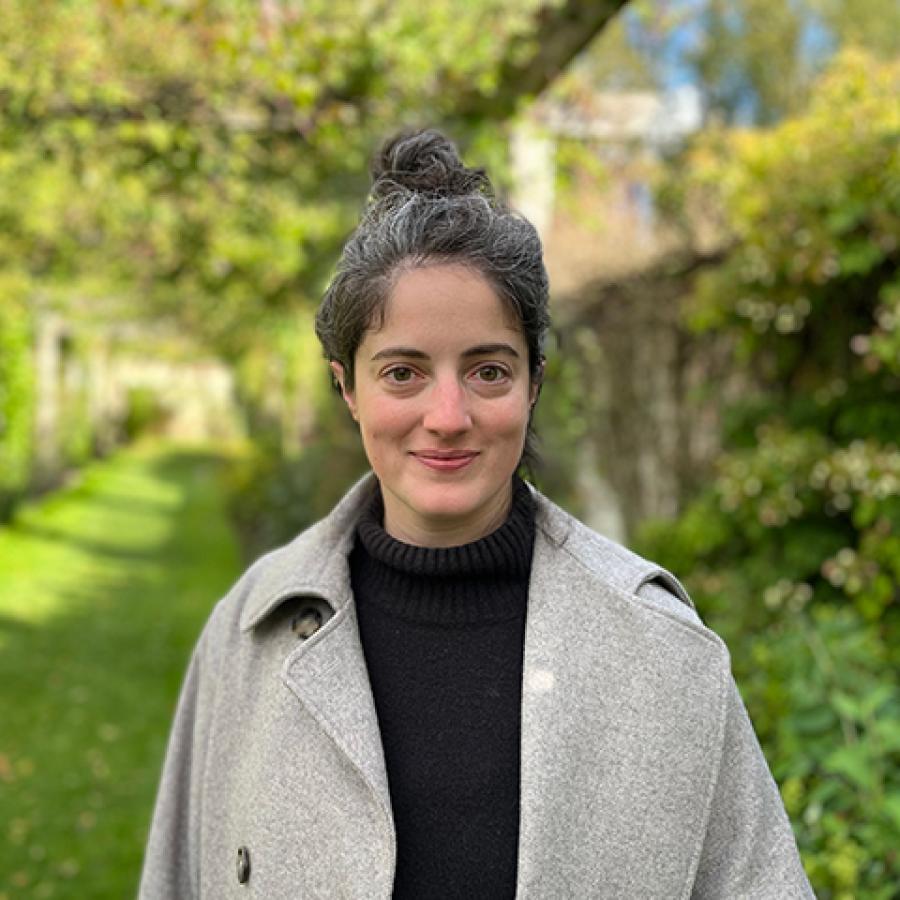 A person with their hair tied up in a bun, wears a black jumper and light grey coat. They're smiling to the camera, as they stand in a garden.