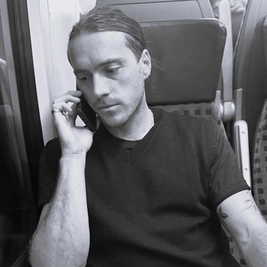 A black and white photo of Ian sitting on a train, holding a phone to his ear.