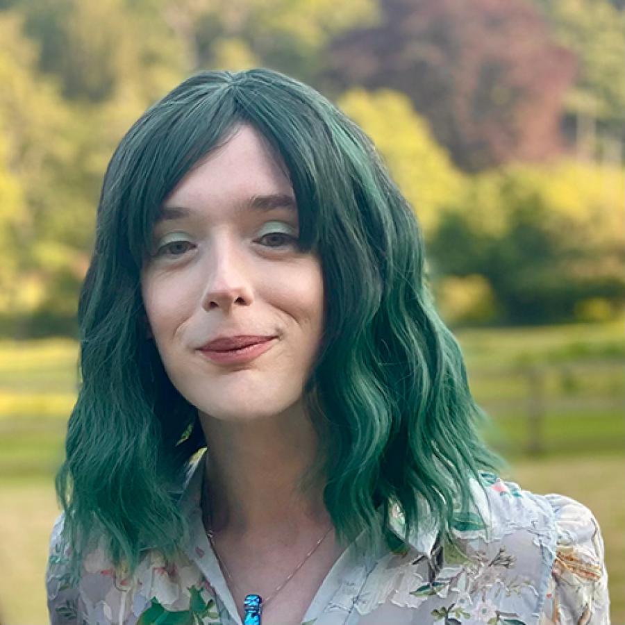 Dr Merlyn Seller has green hair and is wearing a floral blouse. They are standing in a park, smiling to the camera.