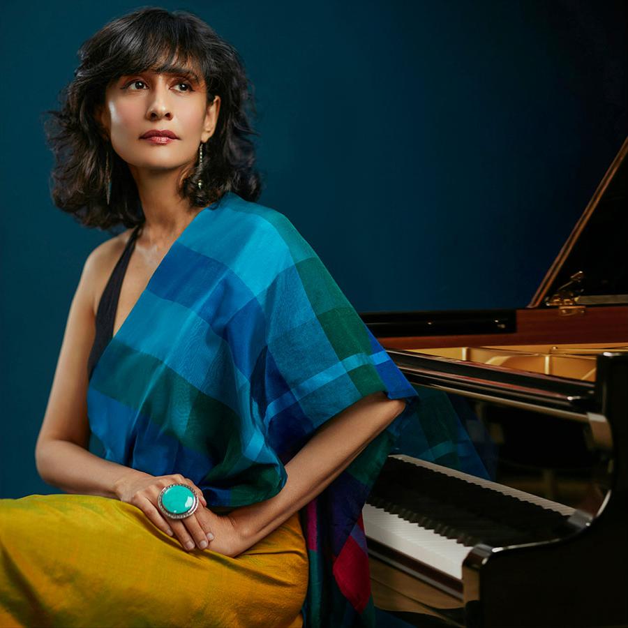 Pianist Dr Tanya Ekanayaka sits by a grand piano. They wear a blue checked shawl and yellow skirt.