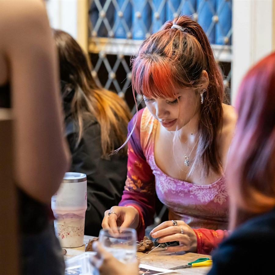 A person with long hair in a pony tail is sitting at a table and working with some clay.