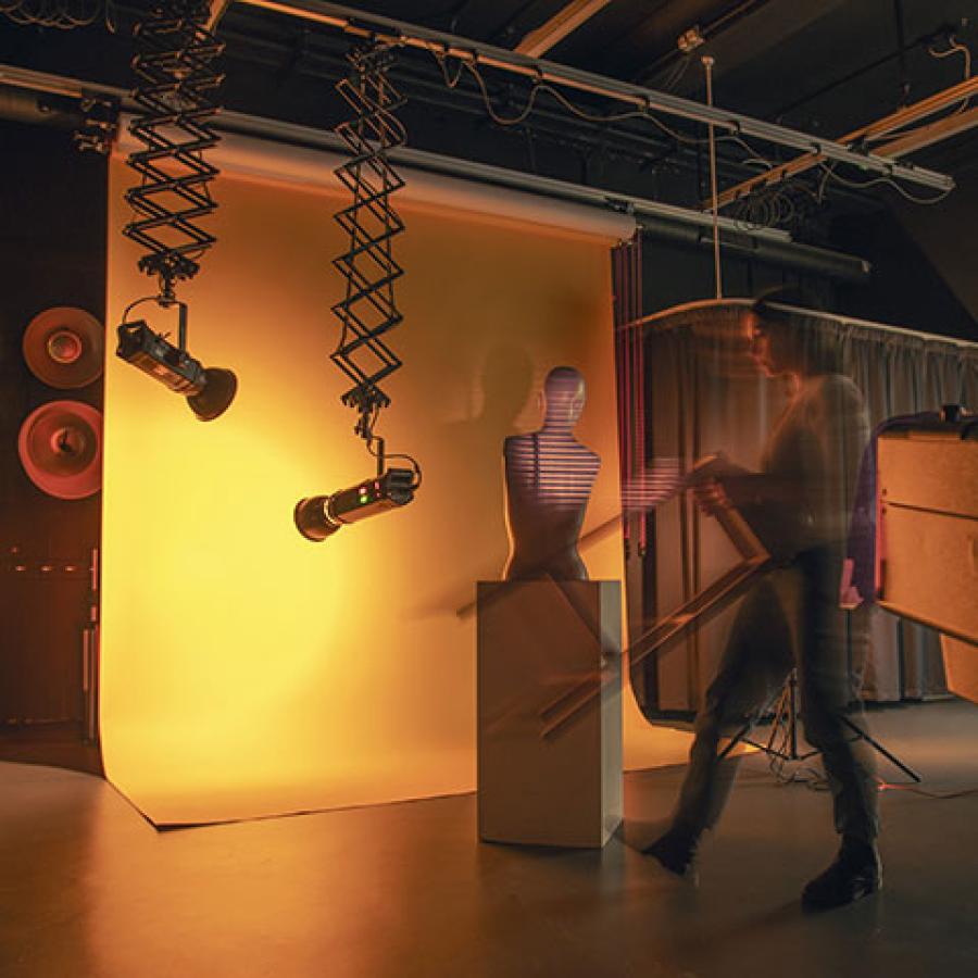 Shareen carrying a stool in the ECA photography studio. There is a screen hanging with a plinth and the top half of a mannequin placed upon it.