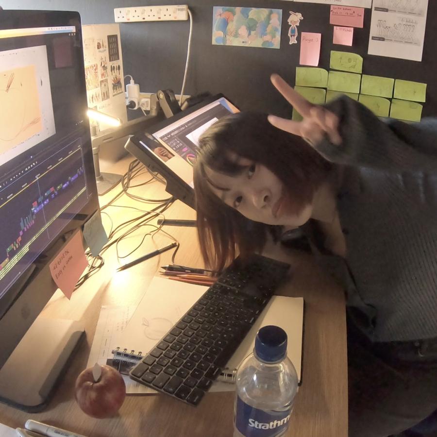 Ruge sits at her desk in front of a large monitor. Her animation work is around her and she does the peace sign to camera.